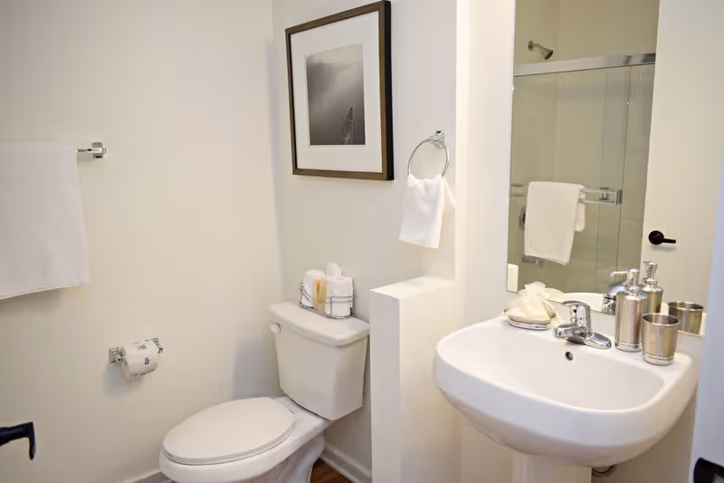 A clean and modern bathroom featuring a white toilet with neatly arranged towels and toiletries on top, a white pedestal sink with silver soap dispensers and a cup, a large mirror above the sink, a towel ring with a white hand towel, a towel bar with a white bath towel, and a framed black and white photo on the wall.