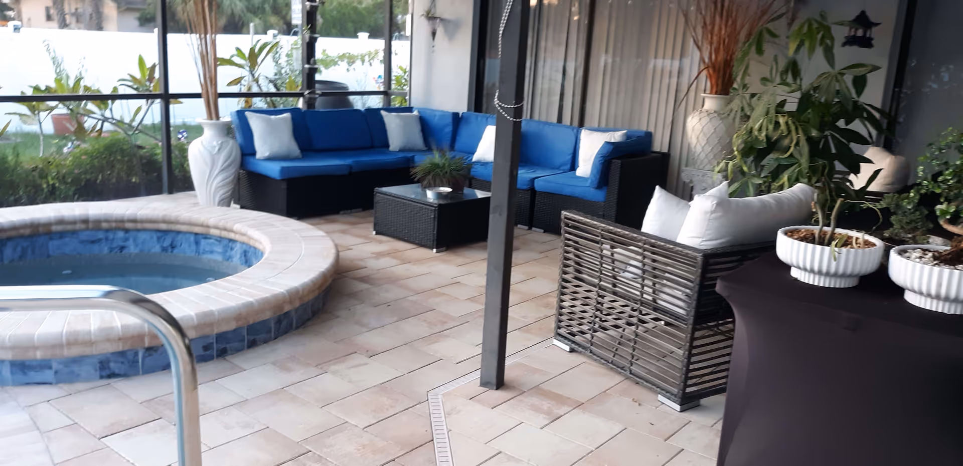 Outdoor patio area with a round hot tub surrounded by tiled flooring. There is a blue cushioned sectional sofa with white pillows, a black wicker armchair with white pillows, a black coffee table with a small plant, and several potted plants around the space. The area is enclosed with glass walls and has a view of greenery outside.