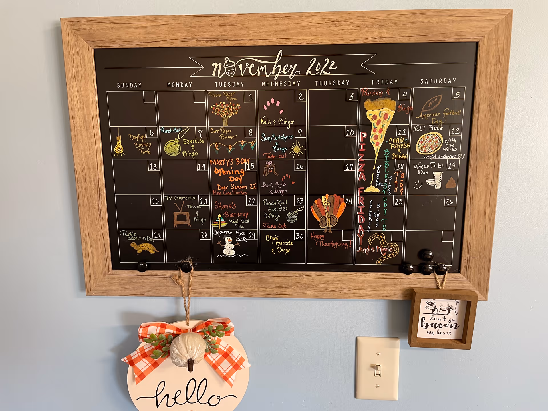 A wooden-framed blackboard calendar for November 2022 with colorful handwritten notes and drawings marking various events and holidays. Below the calendar, a decorative hanging sign with a white pumpkin and a red plaid bow says 'hello'. To the right of the calendar, a small wooden plaque with a pig illustration and the text 'don't go bacon my heart' is hanging on the wall above a light switch.