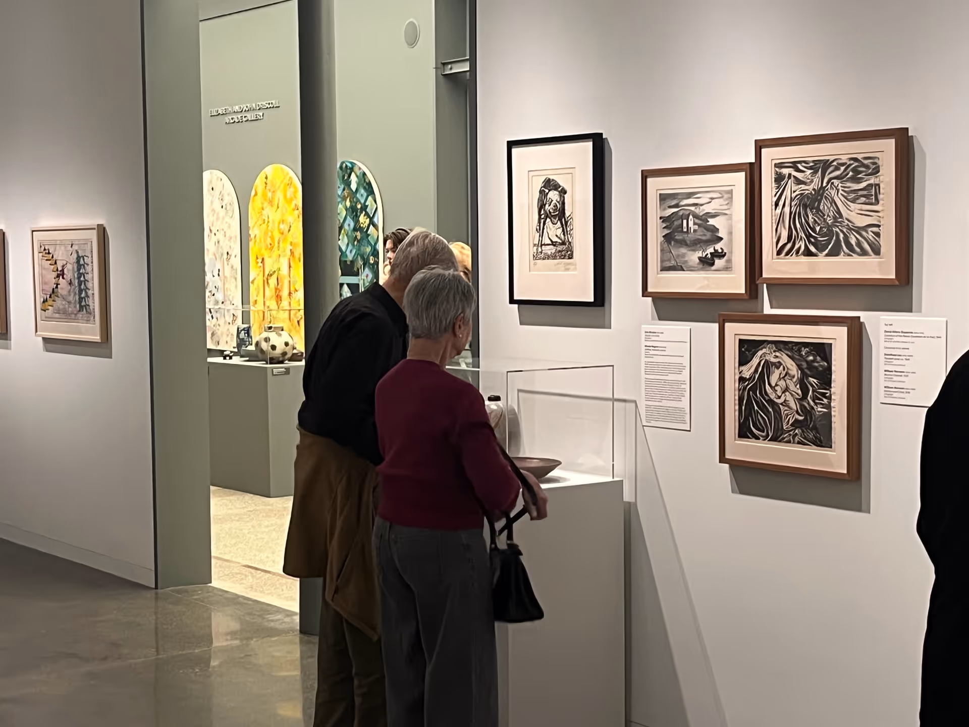 Two elderly people closely observing framed artwork and a display case in an art gallery with various paintings and sculptures visible in the background.