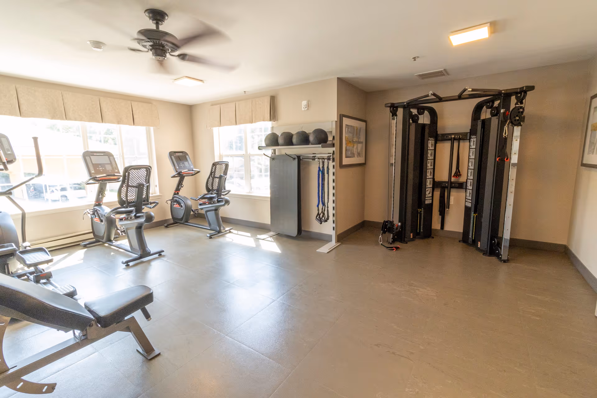 A bright fitness room with exercise equipment including stationary bikes, a weight bench, a multi-functional cable machine, and shelves holding exercise balls and resistance bands. Large windows allow natural light to fill the room.