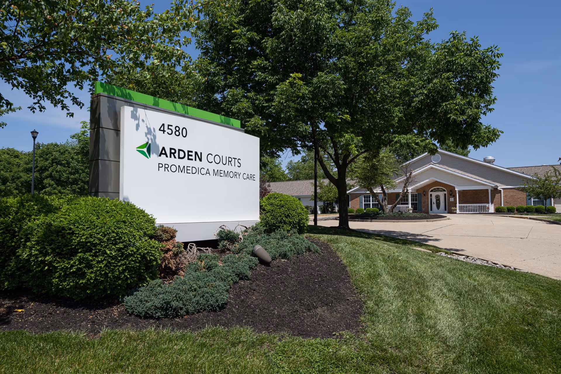 Exterior view of Arden Courts - ProMedica Memory Care Community in Kenwood, showing the facility's sign with the address 4580, surrounded by green bushes and trees, with the building entrance visible in the background under a clear blue sky.