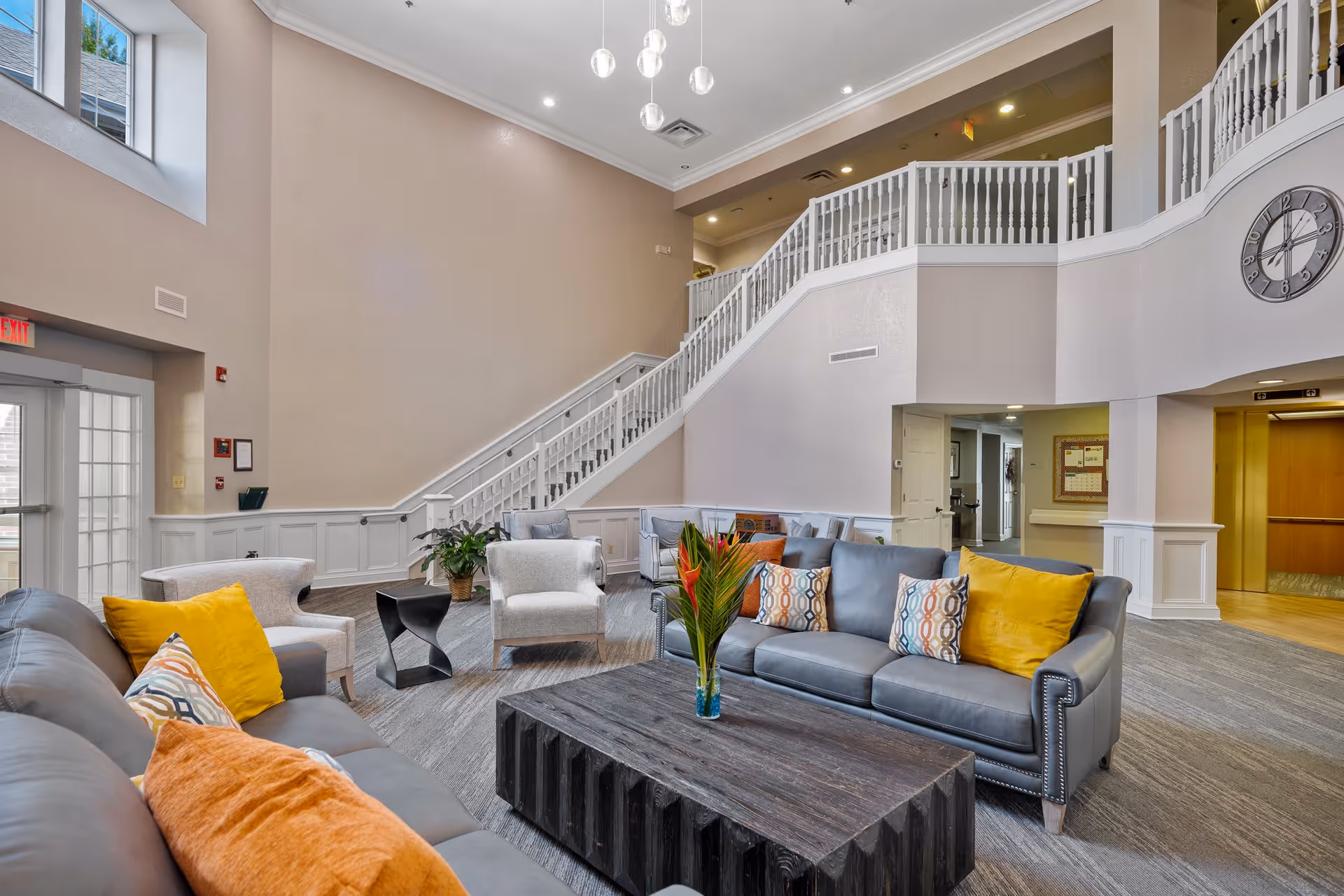 Spacious two-story senior living lounge with sofas, armchairs, a central coffee table, decorative pillows, and a staircase leading to an upper balcony.