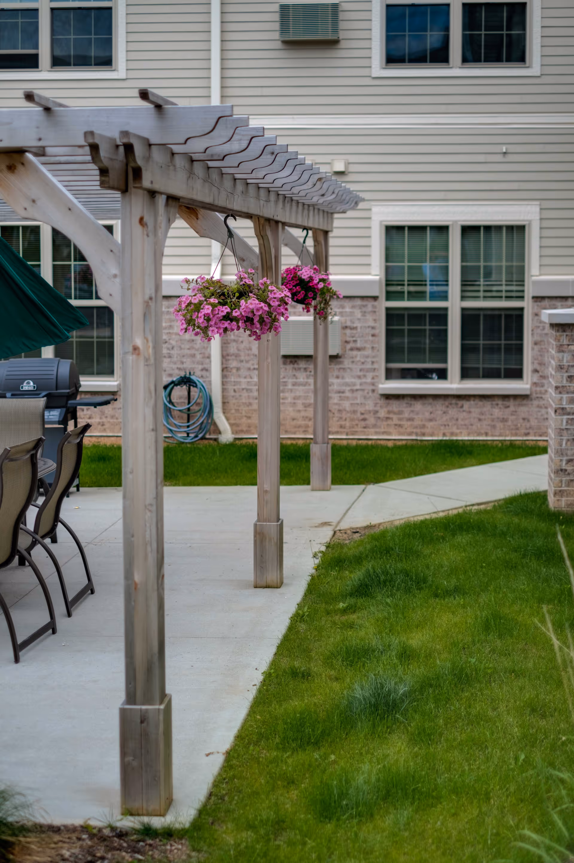 Outdoor patio area at Lincoln Village Senior Campus featuring a wooden pergola with hanging pink and purple flowers, patio chairs, a grill, and a building with beige siding and brick accents in the background.