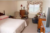 A bedroom in Bethany Village featuring a bed with a light-colored bedspread, a wooden dresser with a lamp, an armchair, a wooden chair, a window with floral curtains, and a TV on a wooden stand.