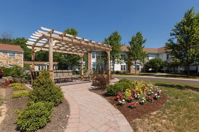 Outdoor garden area at Gwynedd Estates featuring a paved walkway leading to a wooden pergola with benches underneath, surrounded by neatly trimmed bushes, colorful flowers, and trees, with a multi-story building in the background under a clear blue sky.