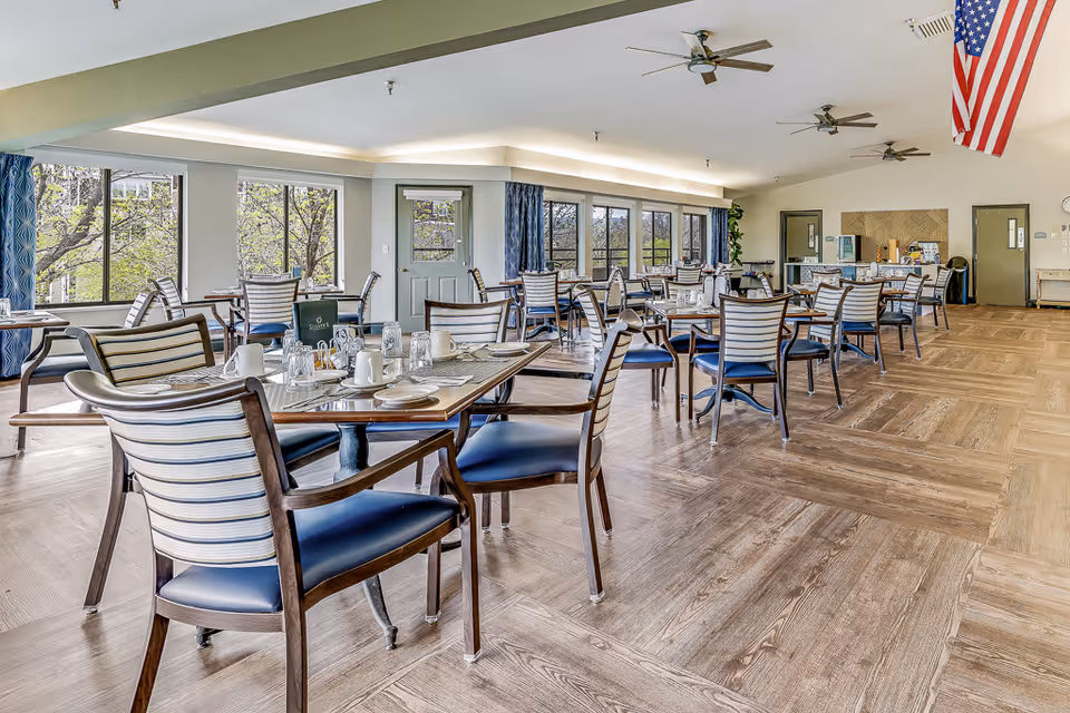 Spacious bright dining room with multiple set tables and striped-back chairs, large windows, and an American flag.