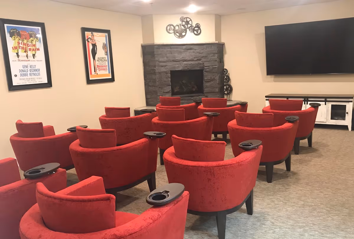 A small screening room with rows of red upholstered chairs facing a fireplace and wall-mounted TV, with movie posters on the wall.