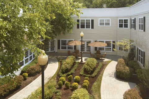 Outdoor courtyard area at Juniper Village at Chatham featuring paved walkways, landscaped garden beds with shrubs and small trees, two patio tables with umbrellas and chairs, and a two-story building with multiple windows surrounding the courtyard.