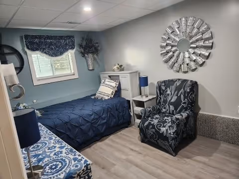 A small, cozy bedroom with a single bed covered in a dark blue comforter and decorative pillows. There is a window with a dark blue valance, a white nightstand with a blue lamp, a white cabinet, and a patterned armchair. The walls are painted light gray and there is a decorative round mirror on the wall.