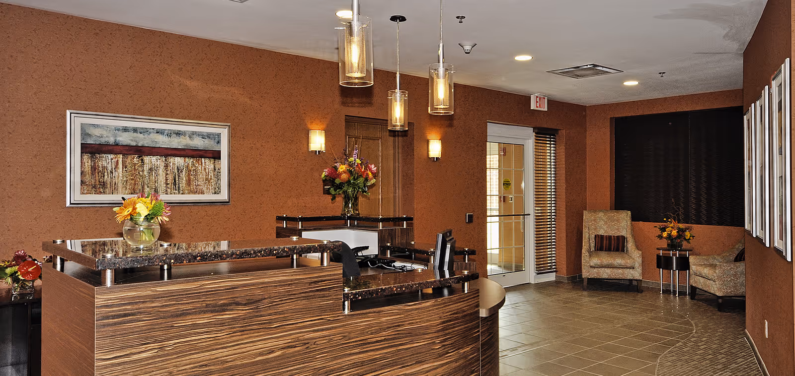 Reception area with a curved wooden desk topped with dark granite, decorated with flower arrangements. The walls are painted a warm brown color and adorned with framed artwork. There are two armchairs with a small table and flowers in the corner near a glass door with blinds. Pendant lights hang from the ceiling, and the floor is tiled.