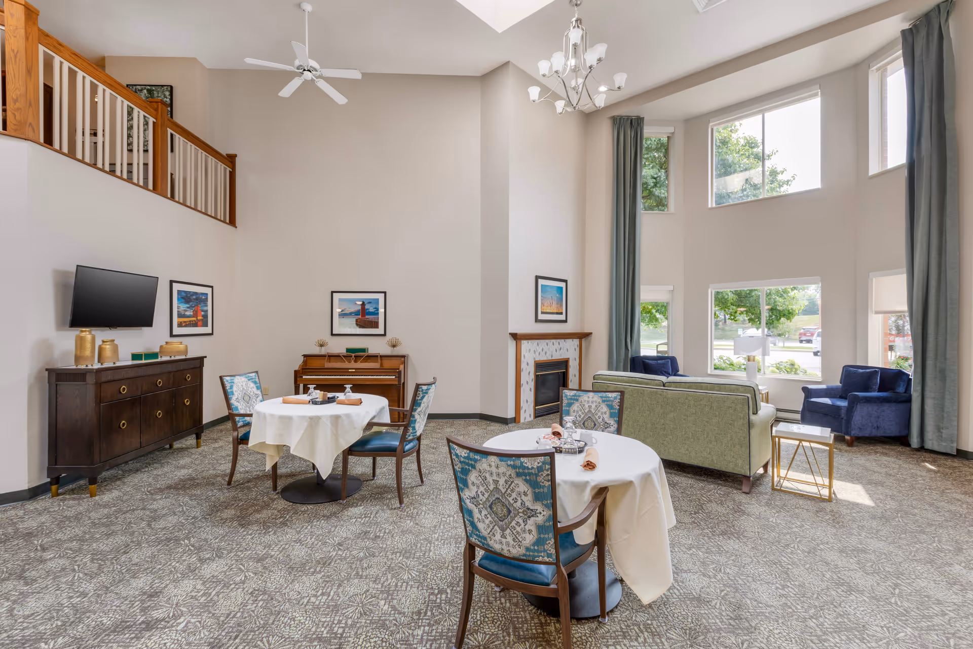 A bright and spacious senior living common area with high ceilings and large windows letting in natural light. The room features two round tables covered with white tablecloths and set with napkins and small trays. There are patterned chairs around the tables. In the background, there is a green sofa, two blue armchairs, a fireplace with a framed picture above it, and a piano against the wall. A ceiling fan and chandelier hang from the ceiling. A flat-screen TV is mounted on the wall above a dark wooden cabinet with decorative items on top.
