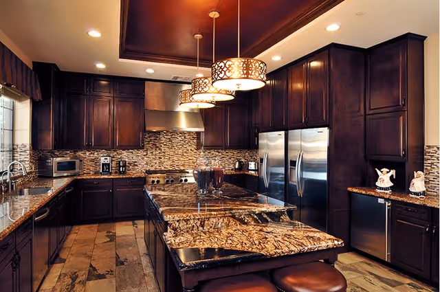 Spacious kitchen with dark wooden cabinets, granite countertops, a large kitchen island with bar stools, stainless steel appliances including two refrigerators and a stove with a range hood, pendant lights hanging above the island, and decorative backsplash tiles.