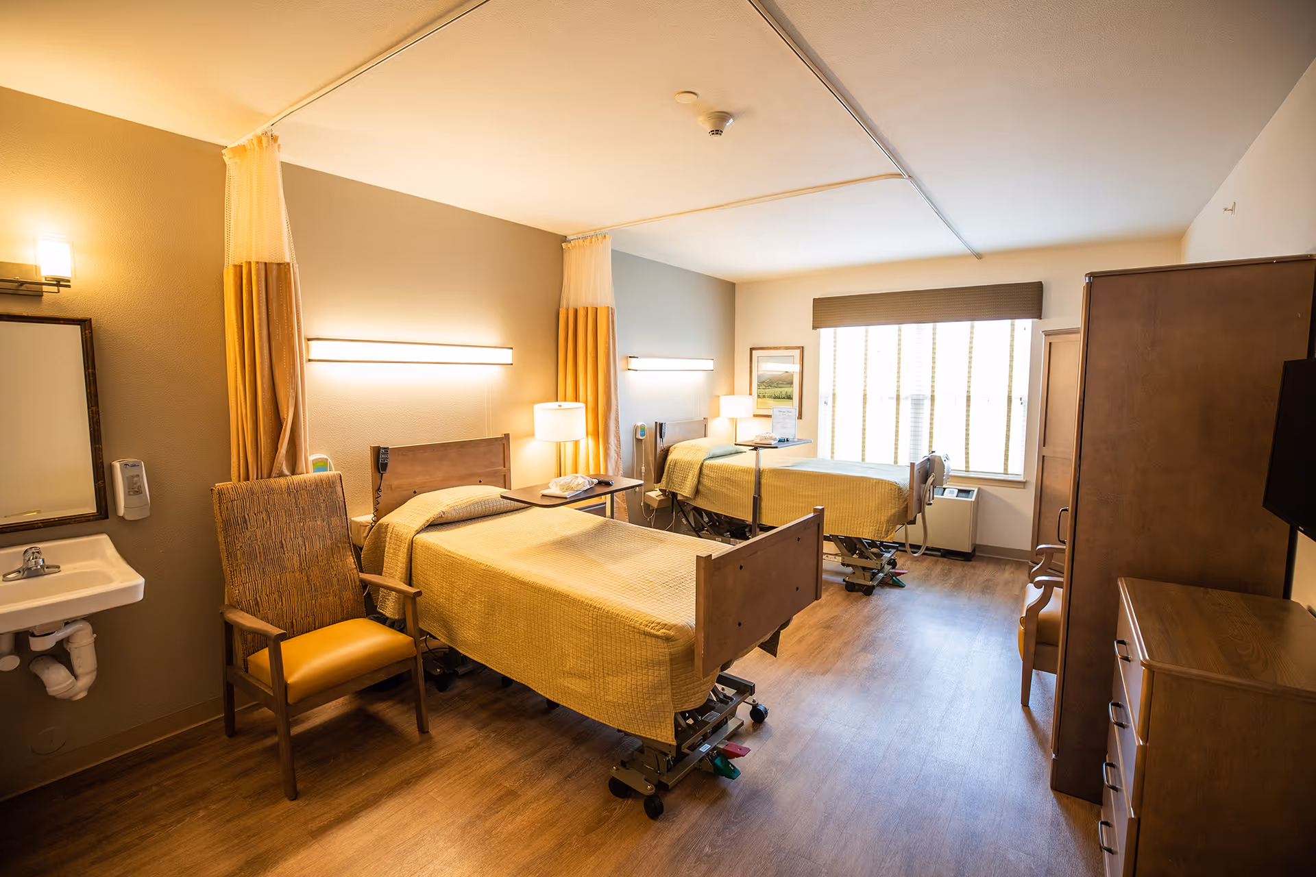 A well-lit room with two hospital-style beds covered in yellow bedspreads, each with a bedside table and lamp. There is a wooden chair with yellow upholstery near a small sink and mirror on the left wall. The room has wooden flooring, a large window with blinds, and wooden furniture including a dresser and a wardrobe.