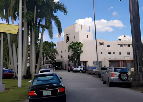Driveway lined with parked cars and palm trees leading to a multi-story beige building under a blue sky.