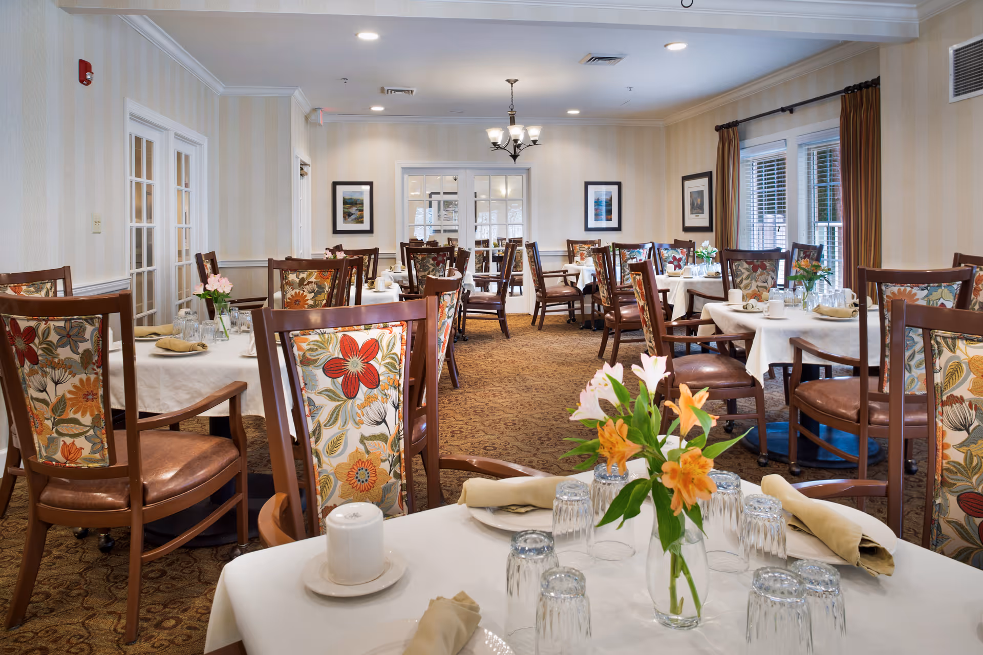 A well-lit dining room with multiple tables covered in white tablecloths, each set with glasses, plates, napkins, and floral centerpieces. The chairs have wooden frames with floral patterned upholstery. The room has beige striped wallpaper, framed artwork on the walls, and windows with brown curtains.
