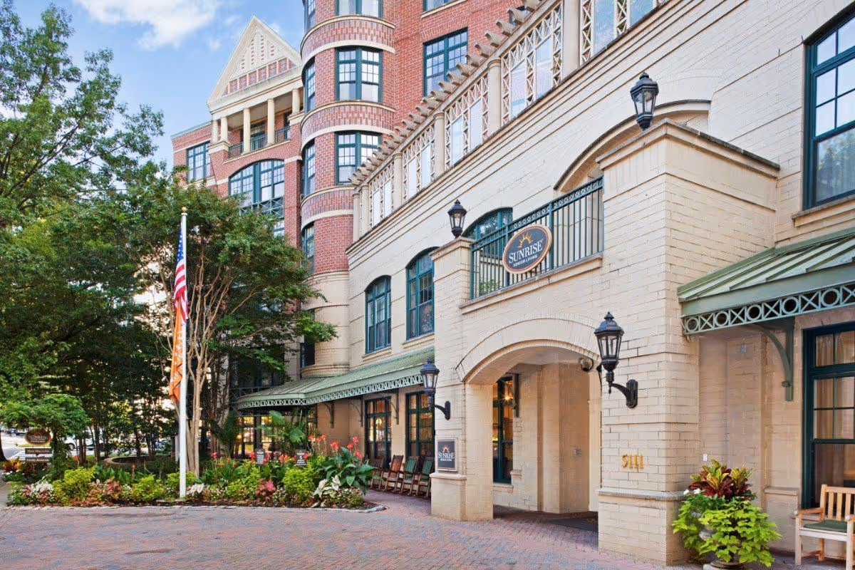 Brick and stone building front with an arched entrance and a 'Sunrise' sign, flagpoles, landscaping, and a covered walkway with seating.