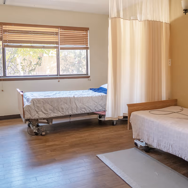 A room with two hospital-style beds separated by a beige privacy curtain. One bed is positioned near a window with wooden blinds, and the floor is covered with wood-like flooring. The room has neutral-colored walls and a small rug on the floor.