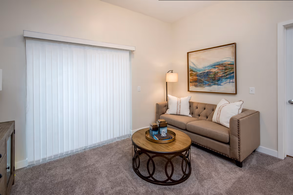 A cozy living room with a beige tufted sofa adorned with two white pillows, a round wooden coffee table with decorative items on top, a floor lamp with a beige shade, a large abstract painting on the wall, and vertical blinds covering a sliding glass door. The room has beige walls and carpeted flooring.
