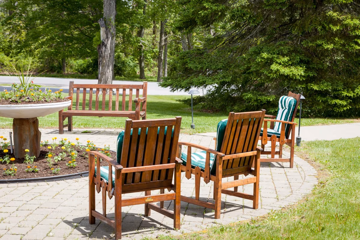 Outdoor seating area with wooden chairs featuring green and white striped cushions arranged on a circular paved patio around a large planter filled with flowers, surrounded by green grass and trees.