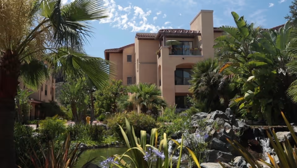 Mediterranean-style apartment building with balconies surrounded by lush tropical landscaping and a small pond.