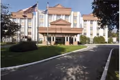Front exterior view of a multi-story nursing and rehabilitation center building with a covered entrance, three flagpoles with flags, and a curved driveway surrounded by grass and trees.