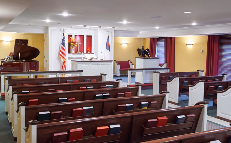 Interior view of a chapel with wooden pews, hymn books, an altar with religious symbols, American and Christian flags, a grand piano, and soft lighting.
