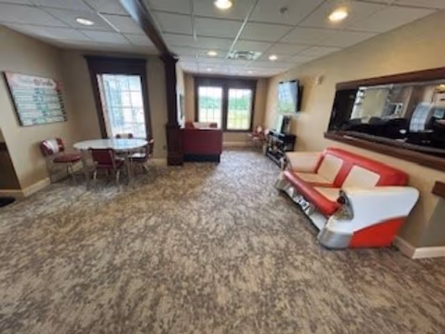 Interior view of a senior living facility common area with a retro-style red and white sofa on the right, a round table with four chairs on the left, a large mirror on the wall above the sofa, a TV mounted on the far wall, and windows letting in natural light.