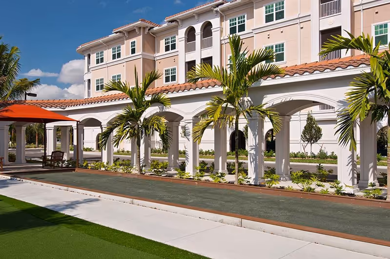 Outdoor view of a senior living facility with a covered walkway featuring white arches and palm trees. There is a bocce ball court in the foreground and a building with multiple windows in the background under a blue sky with some clouds.
