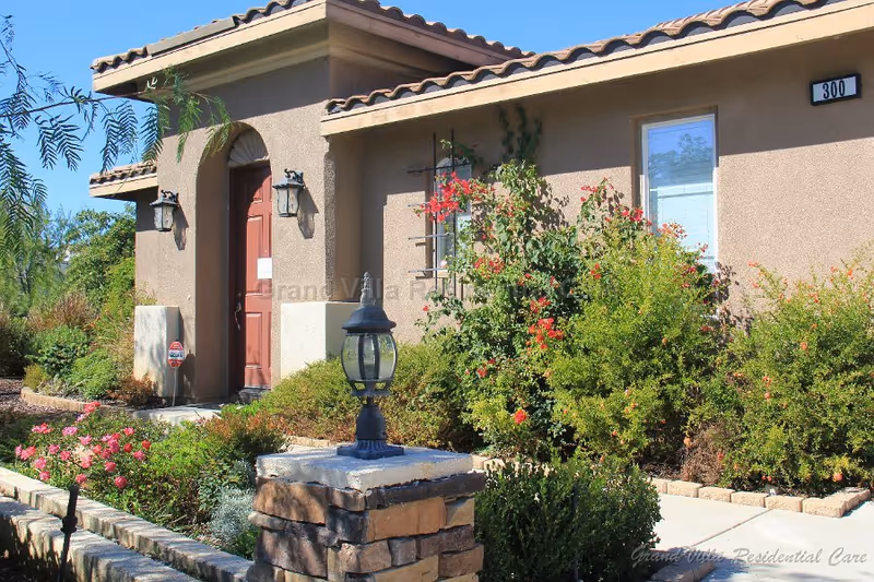 Exterior view of a single-story residential building with a tiled roof and a red front door. The entrance is flanked by two wall-mounted lantern-style lights. There are various green shrubs and flowering plants along the walkway leading to the door. A stone pillar with a black lantern is visible in the foreground. The building number 300 is displayed near a window on the right side.