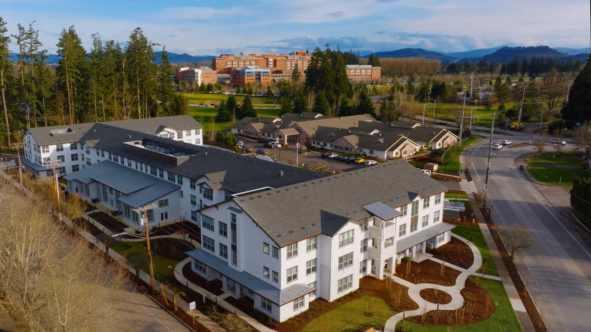 Aerial view of The Esther at Riverbend senior living facility showing multiple connected buildings with gray roofs, surrounded by landscaped grounds, trees, and nearby roads. In the background, there are more buildings and forested hills under a partly cloudy sky.