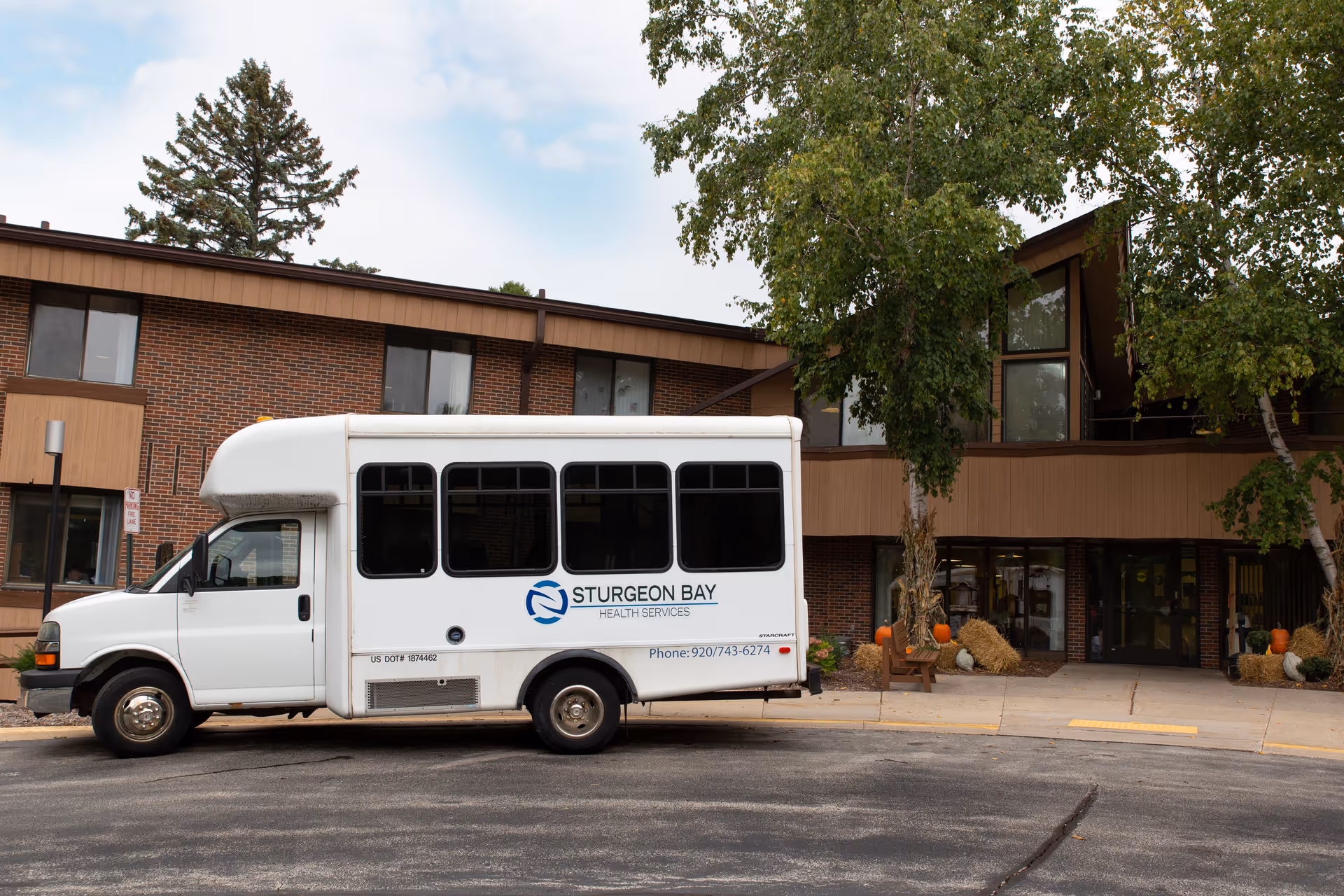 A white shuttle bus with the logo and text 'Sturgeon Bay Health Services' parked in front of a two-story brick building with large windows and trees. The entrance area is decorated with pumpkins and hay bales, suggesting a fall or autumn season.