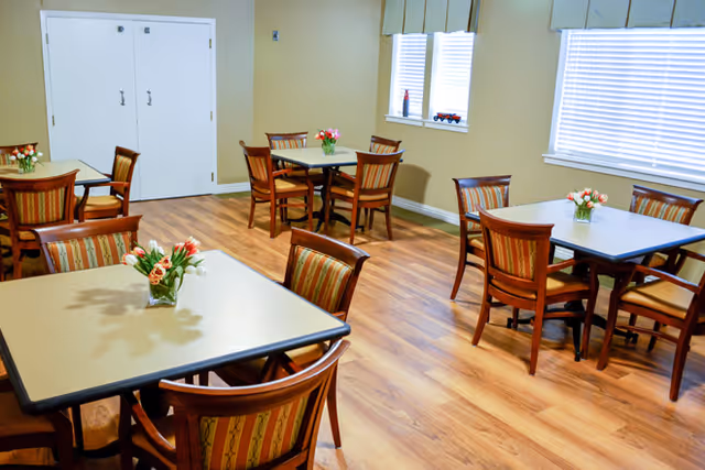 A bright dining room with several square tables, each surrounded by four wooden chairs with striped cushions. Each table has a small vase with flowers. The room has light-colored walls, wood flooring, and windows with white blinds letting in natural light.
