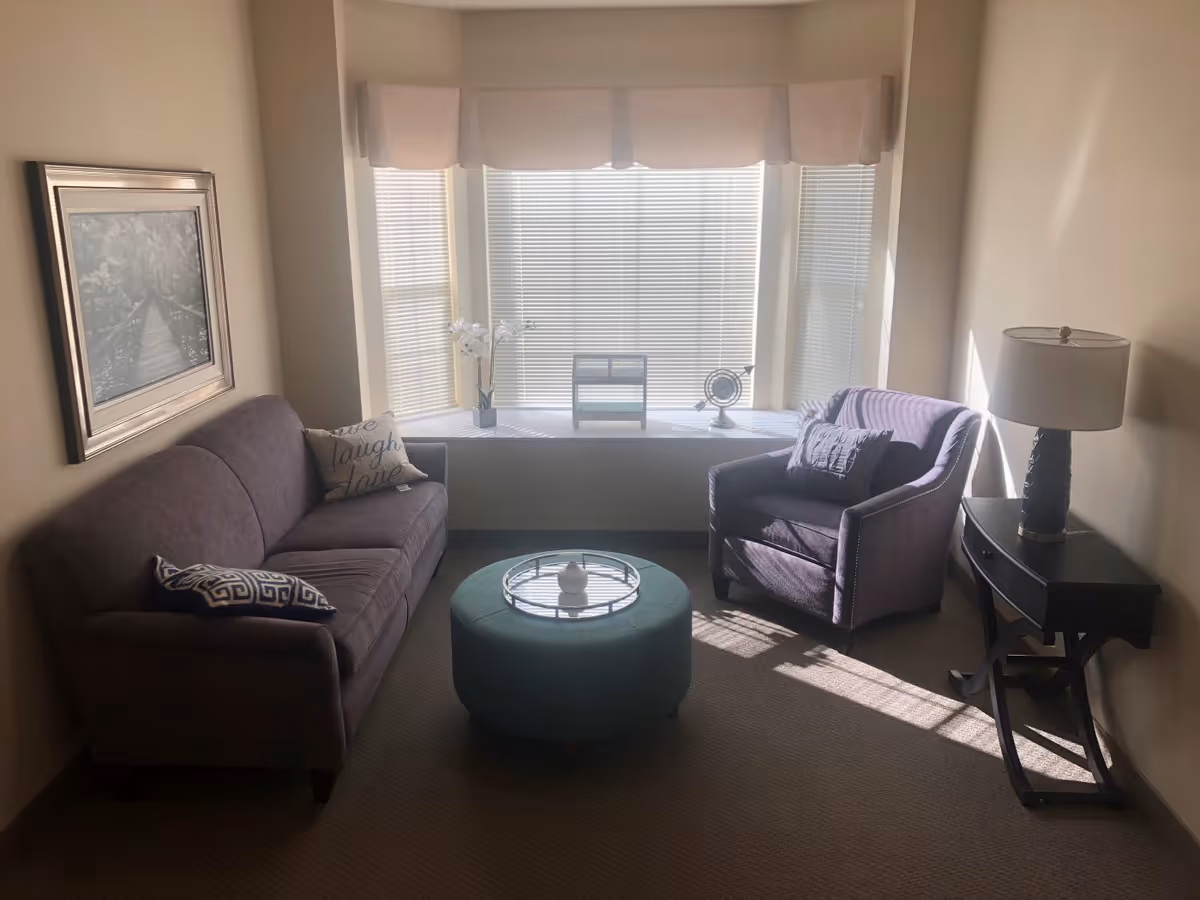 A cozy living room with a purple sofa and matching armchair, a round green ottoman with a decorative tray, a side table with a lamp, and a large window with blinds and a valance. There is a framed picture on the wall and decorative pillows on the sofa.