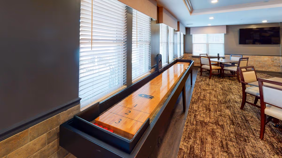 A well-lit common area with a shuffleboard table along the windowed wall and several tables with chairs arranged on a patterned carpet. A flat-screen TV is mounted on the far wall above a brick accent wall.