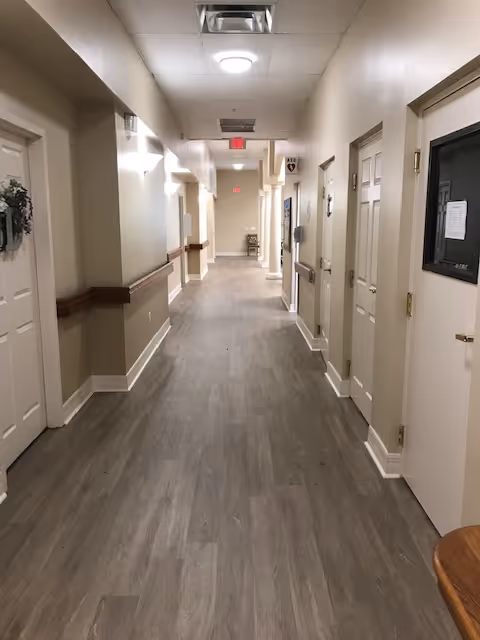 A long, clean hallway in a senior living facility with light-colored walls, wood-like flooring, several closed doors on both sides, handrails along the walls, and ceiling lights. There is an exit sign visible at the far end of the hallway.