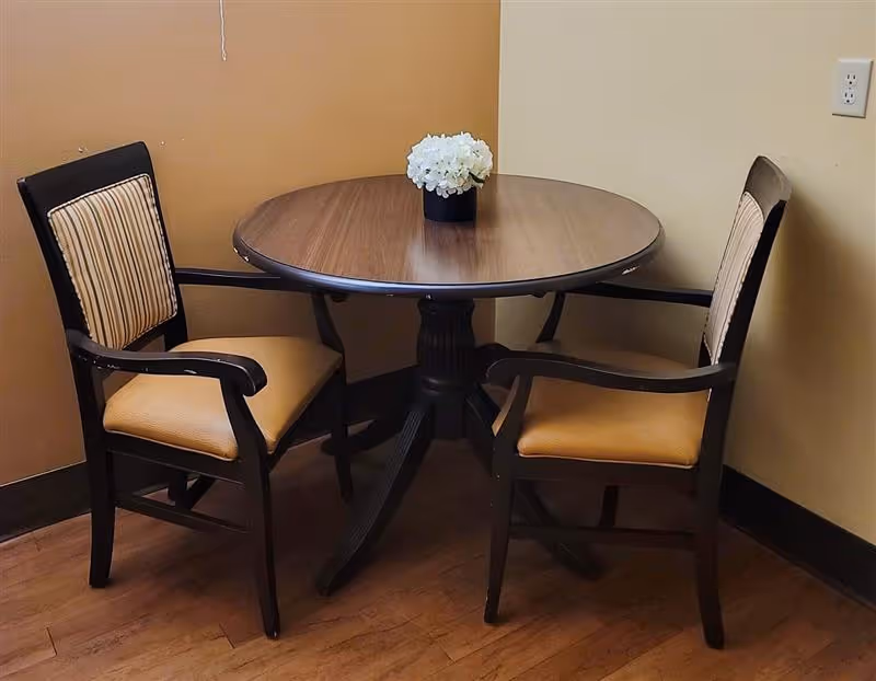 A small round wooden table with a black pedestal base, surrounded by two wooden chairs with beige cushioned seats and striped backrests, placed in the corner of a room with tan and cream-colored walls. A small black vase with white flowers is centered on the table.