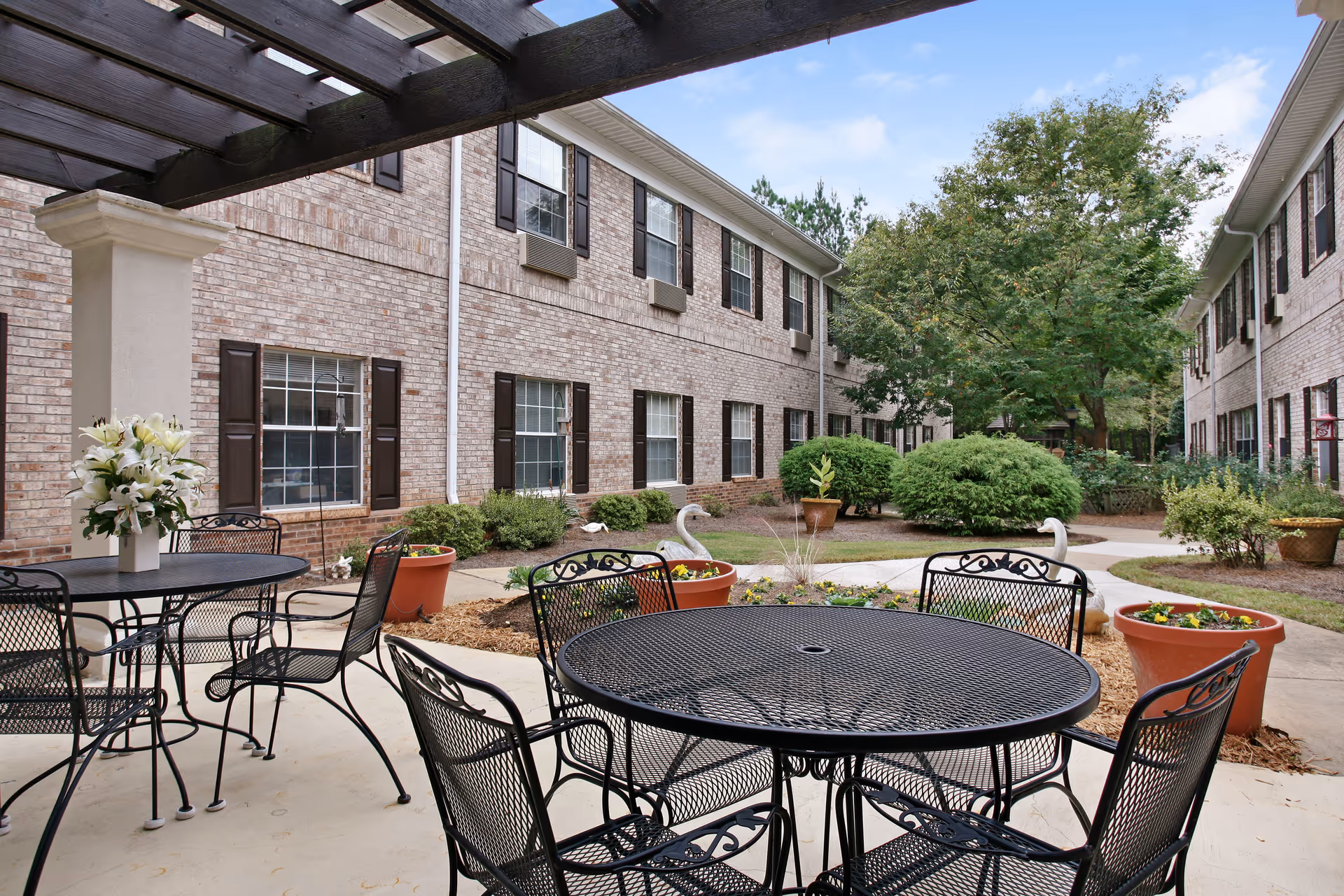 Courtyard with metal patio tables and chairs under a pergola between two-story brick senior living buildings, with potted plants and landscaping.