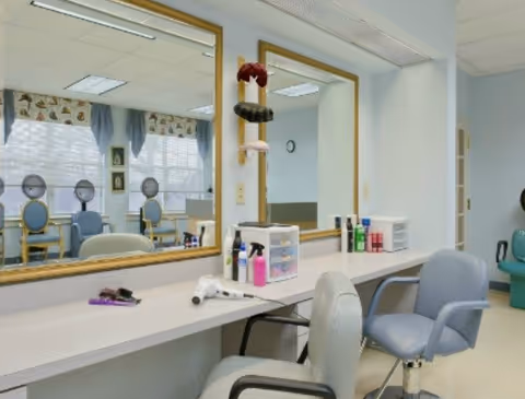 Interior view of a salon area in a senior living facility with two salon chairs in front of a long counter with mirrors. Various hair care products and tools are placed on the counter. In the background, there are additional chairs and windows with curtains.