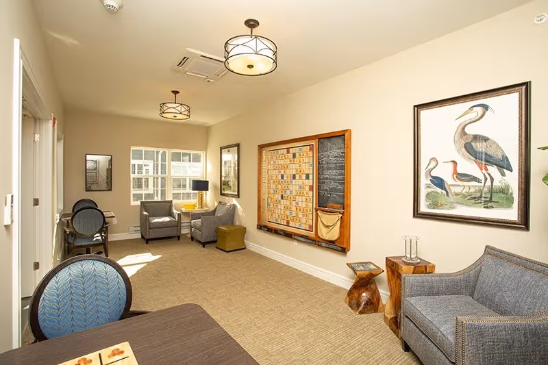 Bright senior living common room with armchairs, a table, a large wall-mounted game board, and framed bird artwork.