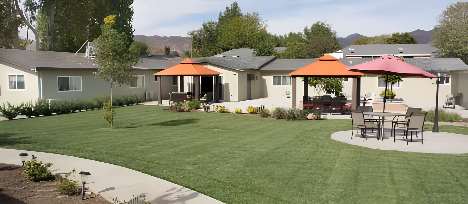 Outdoor area of a senior living facility with green lawn, trees, and several patio seating areas with umbrellas. The building is single-story with light-colored walls and multiple windows. Mountains are visible in the background under a partly cloudy sky.
