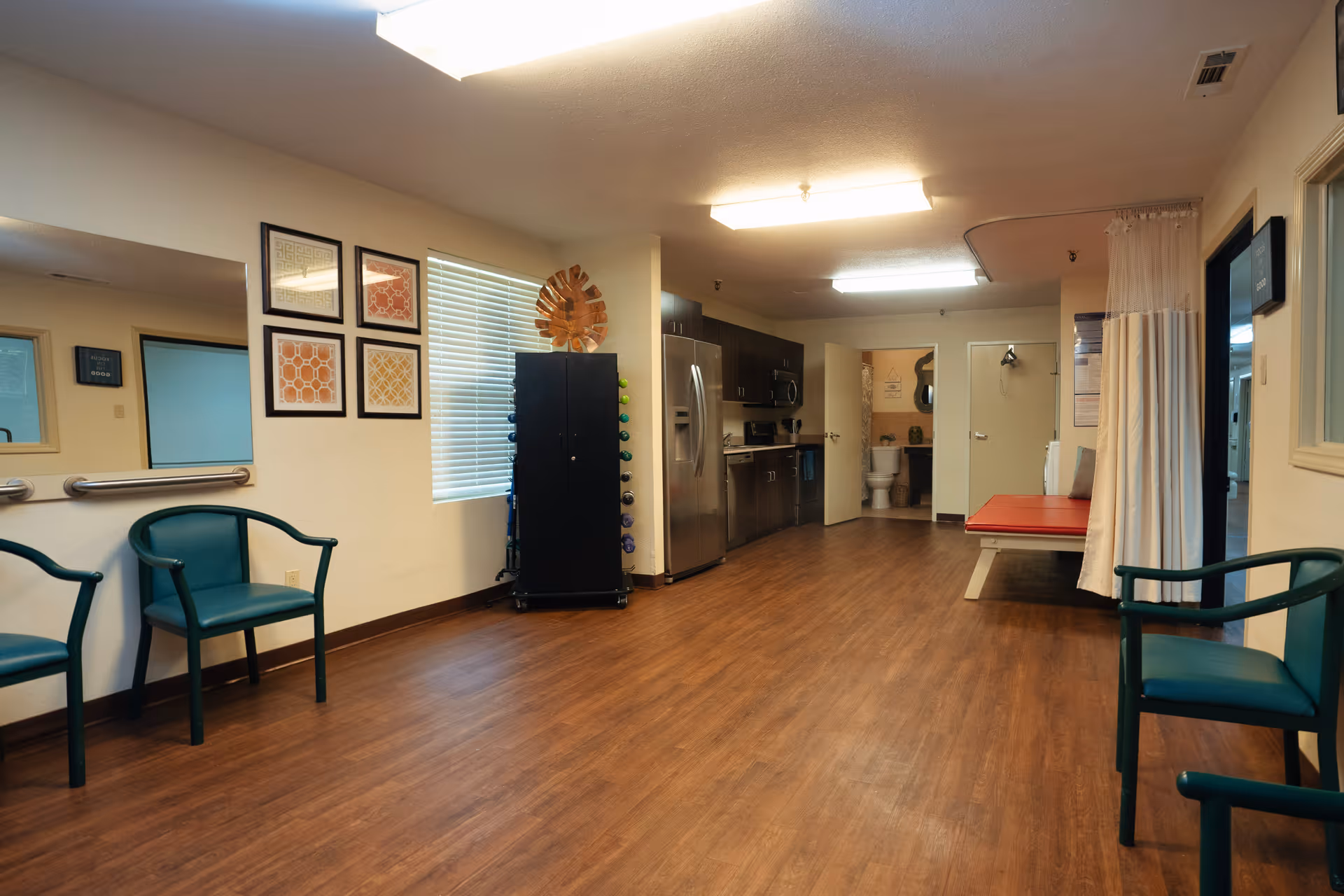 Spacious nursing facility interior with wood floors, green chairs, a kitchenette, a rack of hand weights, and a treatment table by a curtained area.