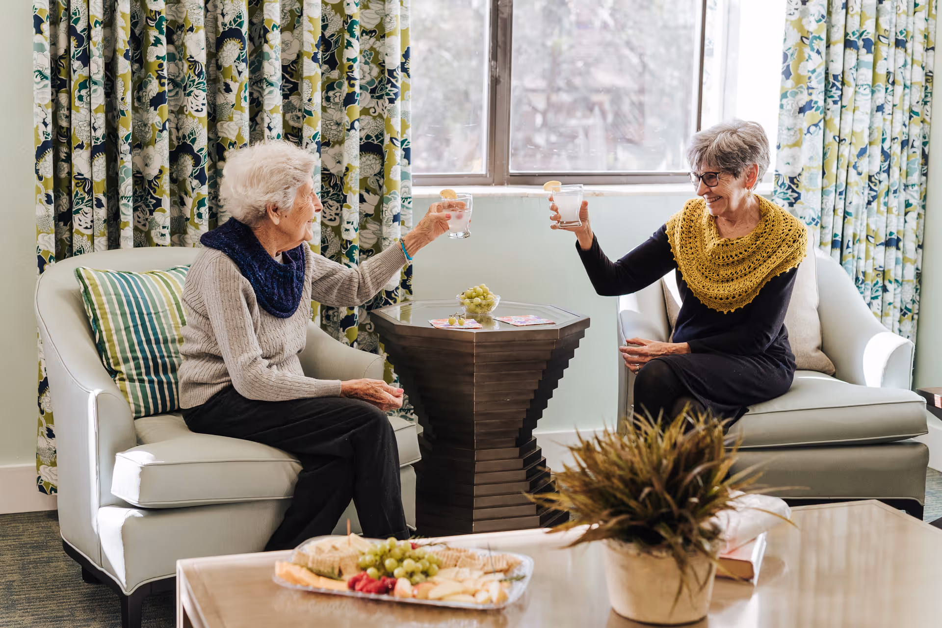 Two elderly women sitting on armchairs in a cozy living room with floral curtains, clinking glasses of water with lemon slices. A small wooden table with grapes and magazines is between them, and a coffee table with a plant and a tray of snacks is in the foreground.