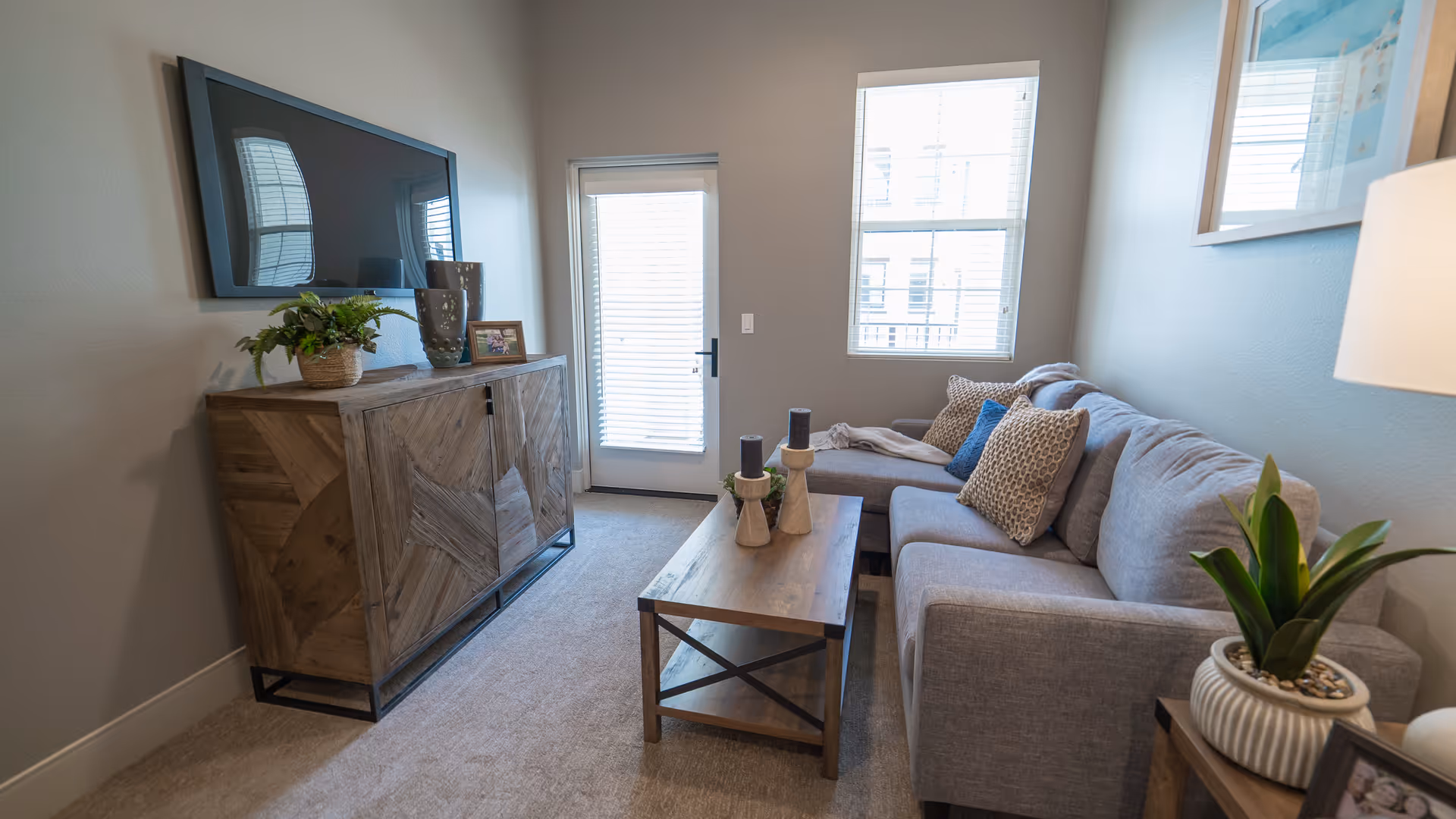 A cozy living room with a gray sectional sofa adorned with patterned and solid pillows. A wooden coffee table with decorative candles is in front of the sofa. Across from the sofa, a wooden cabinet holds a plant, vases, and a framed photo, with a flat-screen TV mounted on the wall above it. The room has a window and a glass door with blinds, allowing natural light to enter. A framed picture hangs on the wall above a side table with a potted plant and a lamp.