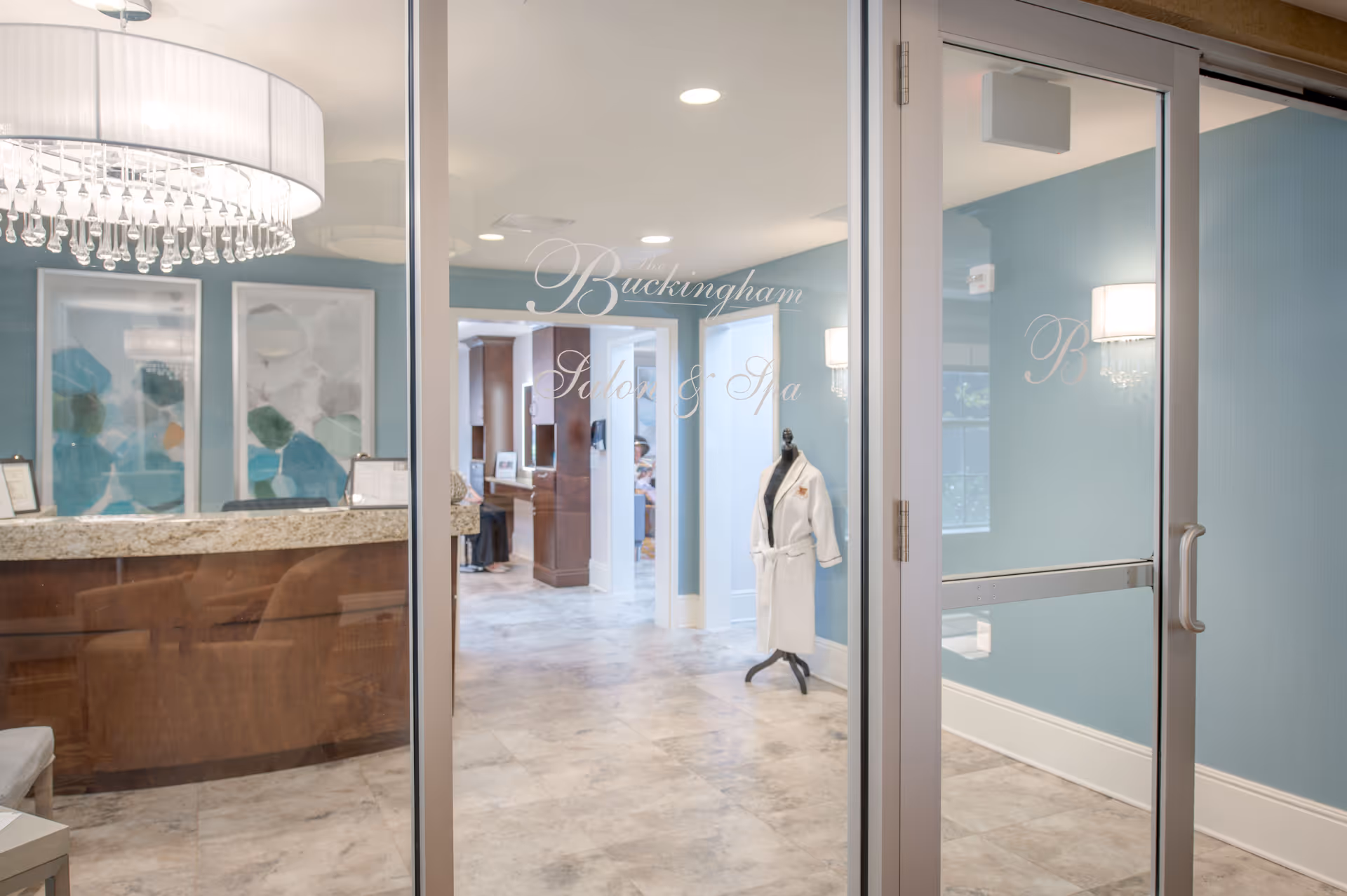 Interior view of The Buckingham Salon & Spa entrance with glass doors, a reception desk with granite countertop, blue walls, framed abstract artwork, a chandelier, and a mannequin wearing a white robe.