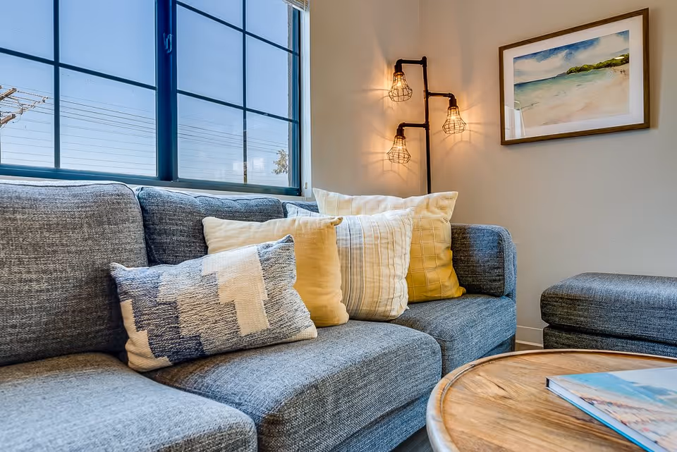 A cozy living room area with a gray fabric sofa adorned with multiple patterned and solid-colored pillows. Next to the sofa is a round wooden coffee table with a book on it. On the wall above the sofa is a framed painting of a beach scene, and a unique industrial-style wall lamp with three exposed bulbs is mounted nearby. A large window with black grid frames lets in natural light.