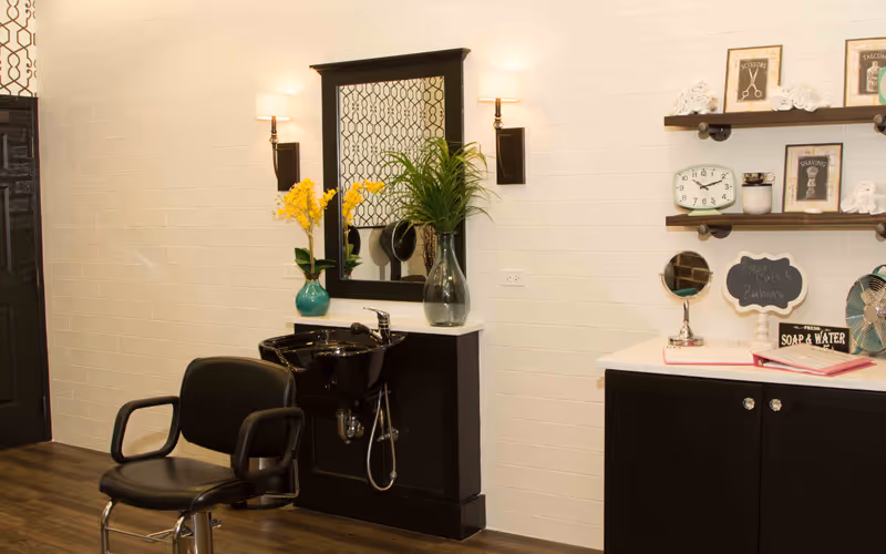 Interior view of a salon area in a senior living facility with a black salon chair in front of a black sink and a large mirror mounted on the wall. The wall is white with two wall sconces on either side of the mirror. There are decorative plants in vases on the counter. On the right side, there are shelves with framed pictures, rolled towels, a clock, and a small chalkboard sign that says 'Fresh Towels'.