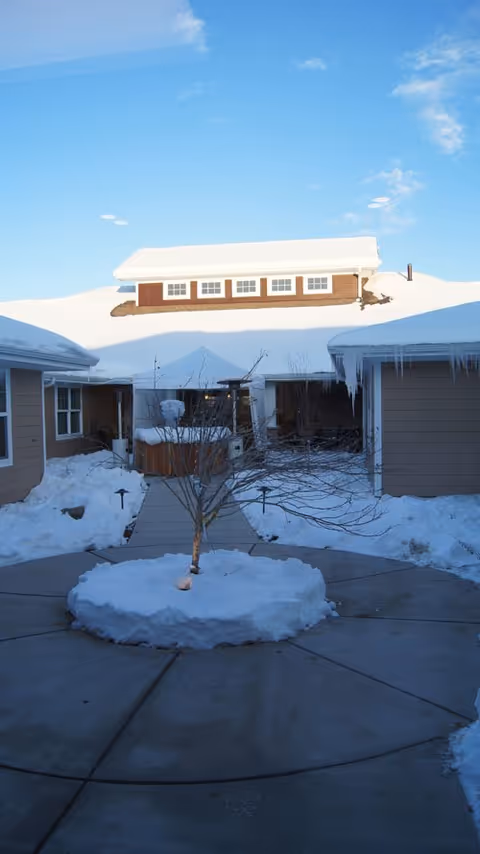 Snow-covered courtyard with a small leafless tree in the center surrounded by a circular concrete path. The courtyard is flanked by buildings with snow and icicles on the roofs. In the background, a snow-covered hill with a building on top is visible under a clear blue sky.