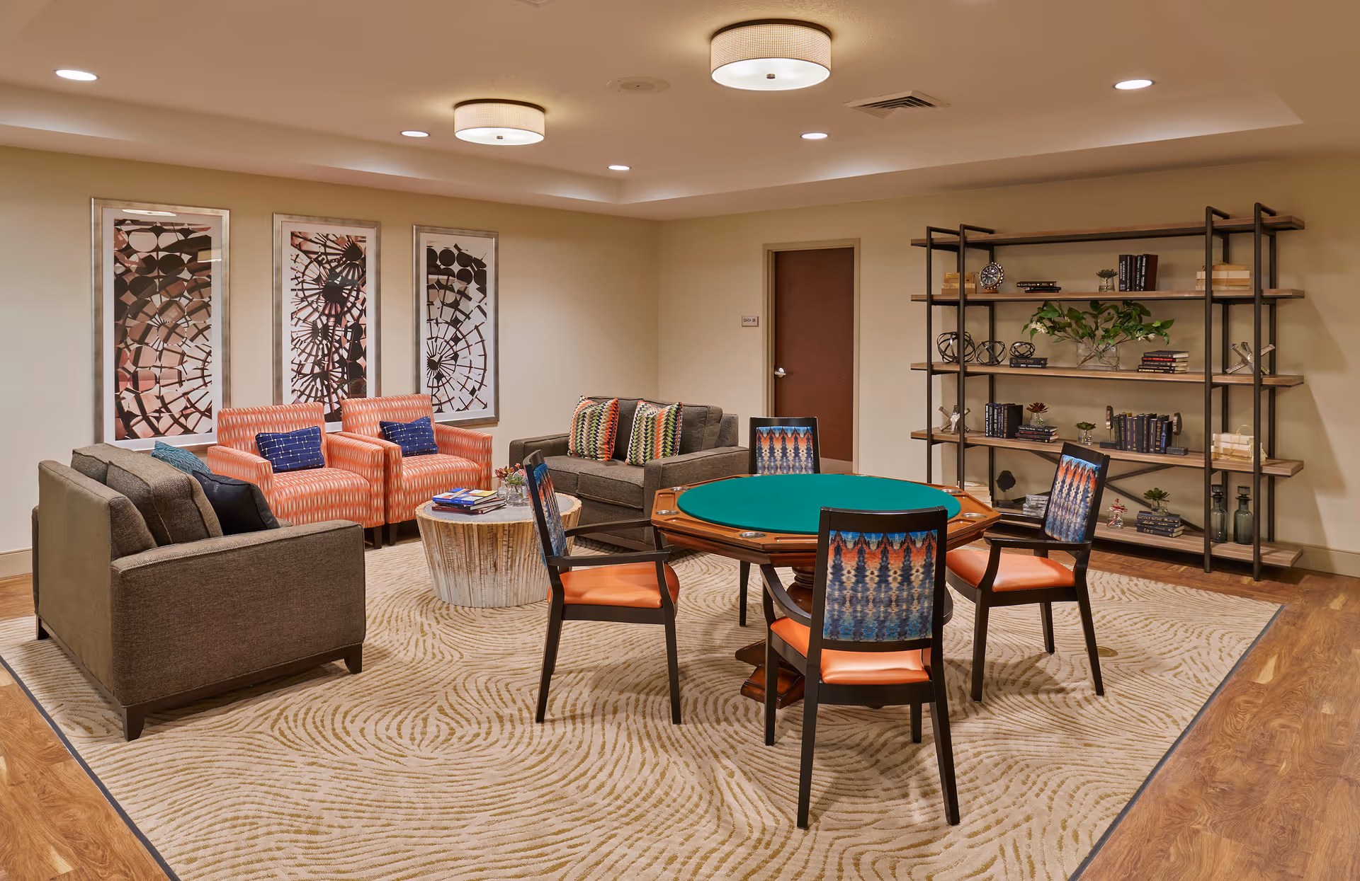 Communal lounge with a green-felt card table and chairs, sofas and armchairs, an area rug, wall art, and a shelving unit.