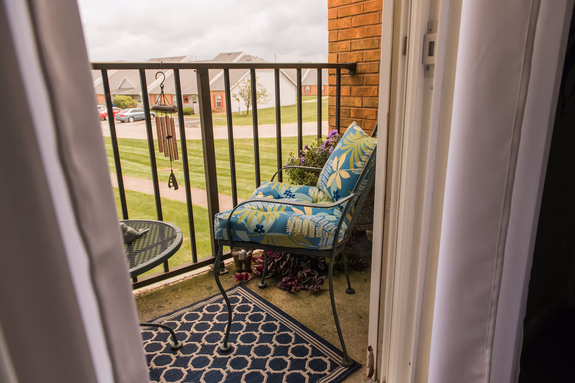 View through an open doorway onto a small balcony with a metal chair featuring blue and green floral cushions, a small round metal table, a hanging wind chime, and a patterned outdoor rug. In the background, there is a grassy area with a sidewalk and residential buildings under a cloudy sky.